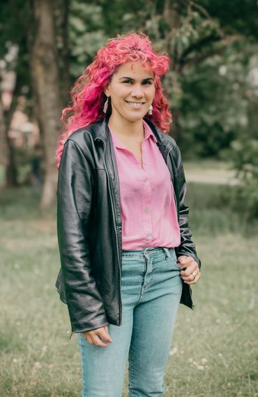 Smiling woman with vibrant pink hair wearing a black leather jacket outdoors.