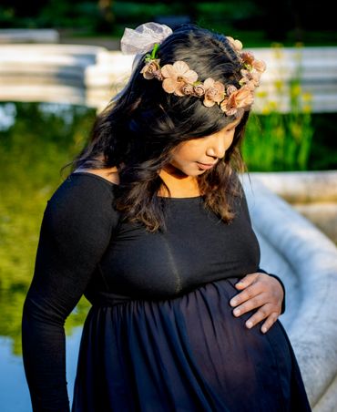 Pregnant woman in black dress with floral crown gently touching her belly outdoors.
