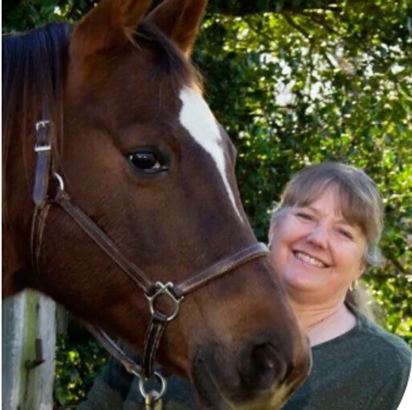 A smiling woman stands next to a brown horse with a white stripe on its face.