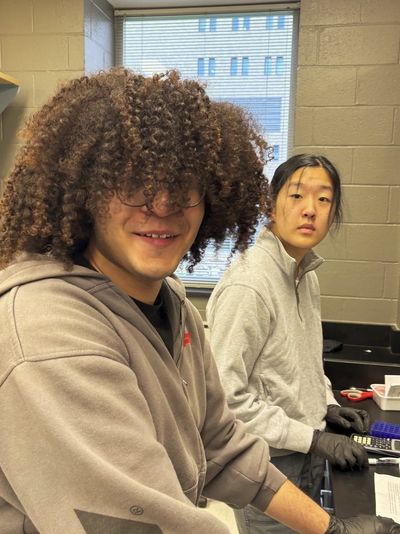 Two young people in a lab setting, one smiling with curly hair, the other focused.