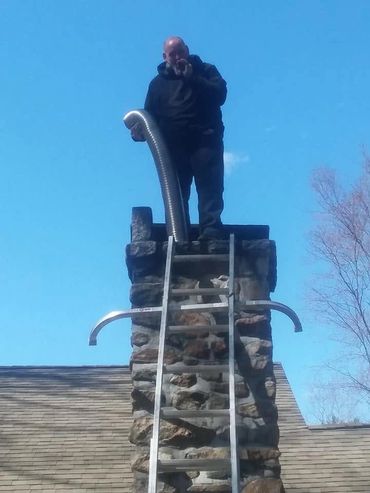 Man standing on a chimney holding a flexible metal tube against a clear blue sky.