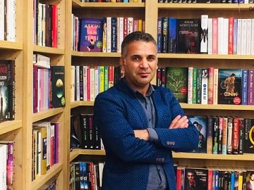 A man in a blue jacket stands confidently in a room filled with bookshelves.