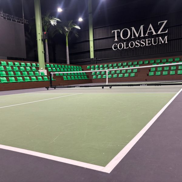 Indoor tennis court with green seating and 'TOMAZ COLOSSEUM' signage.