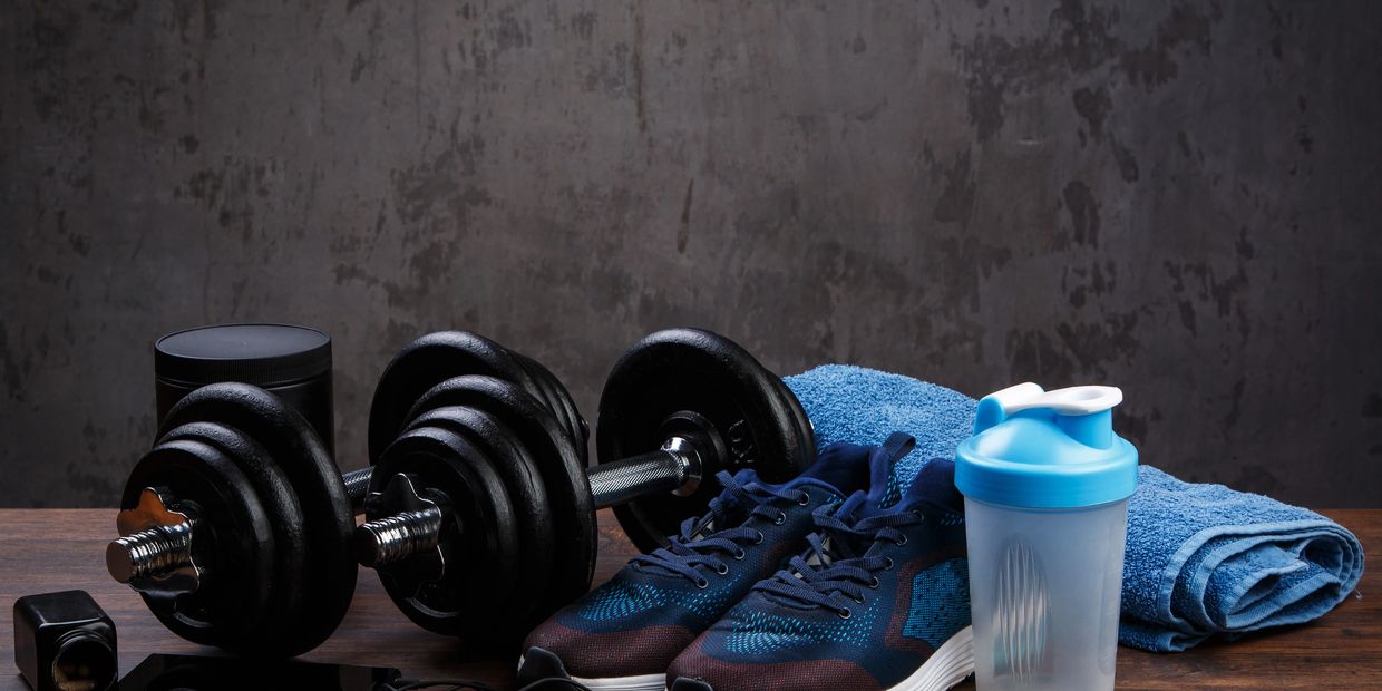 Dumbbells on a table, athletic shoes, a towel, headphones, vitamins, a shaker bottle and pre-workout