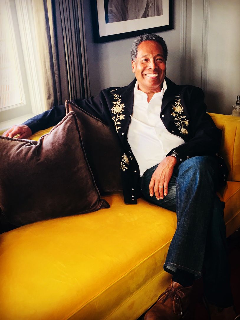 A man in a black embroidered jacket smiles while sitting on a yellow couch.