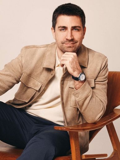 Man in beige jacket sitting on a wooden chair, smiling thoughtfully.