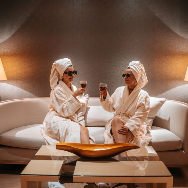Two women in bathrobes and towels on their heads toast with drinks in a cozy lounge.