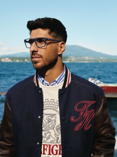 Man wearing glasses and a varsity jacket by the water with mountains in the background.