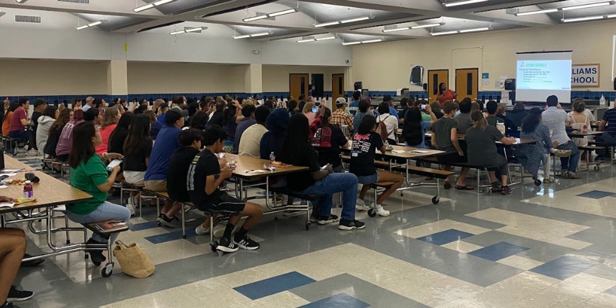 A large group attends a presentation in a school cafeteria.