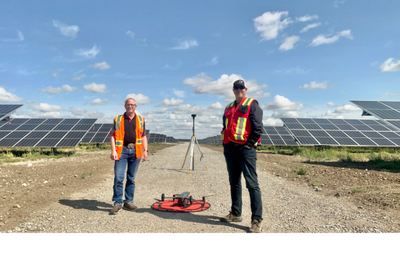 Two workers stand between rows of solar panels with a drone and surveying equipment outdoors.