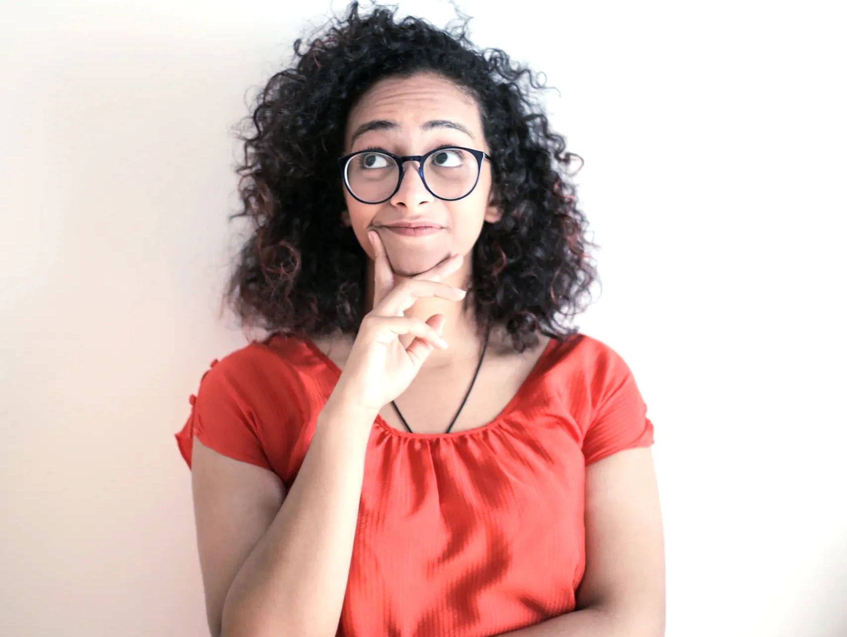 Woman in Red Top Wearing Black Framed Eyeglasses Standing In Front of White Background Thinking