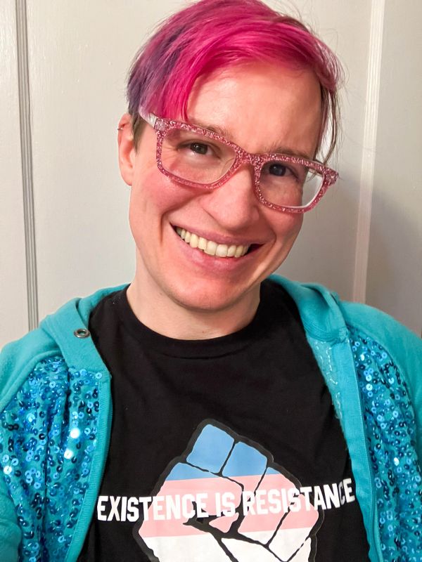 Person with pink hair and glasses smiling, wearing a shirt.