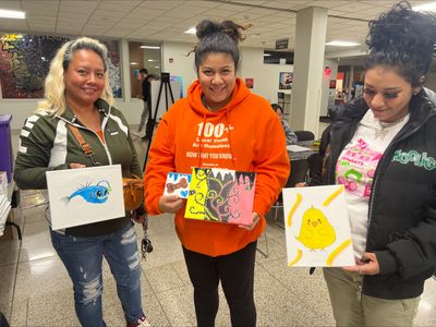 Three women proudly display their colorful paintings indoors.