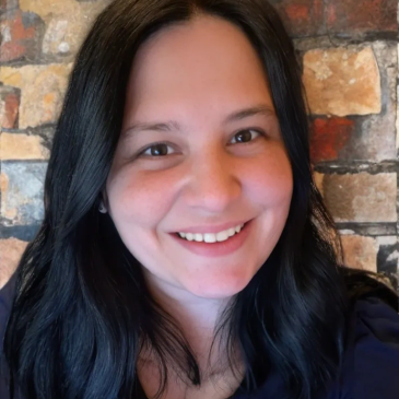Smiling woman with long black hair and a brick wall background.