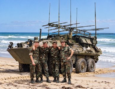 Four soldiers in camouflage pose on a beach with a military vehicle.