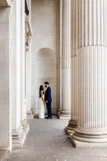 Couple in wedding attire standing close between large classical columns.