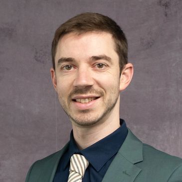 A man in a green suit jacket and striped tie smiles against a gray backdrop.