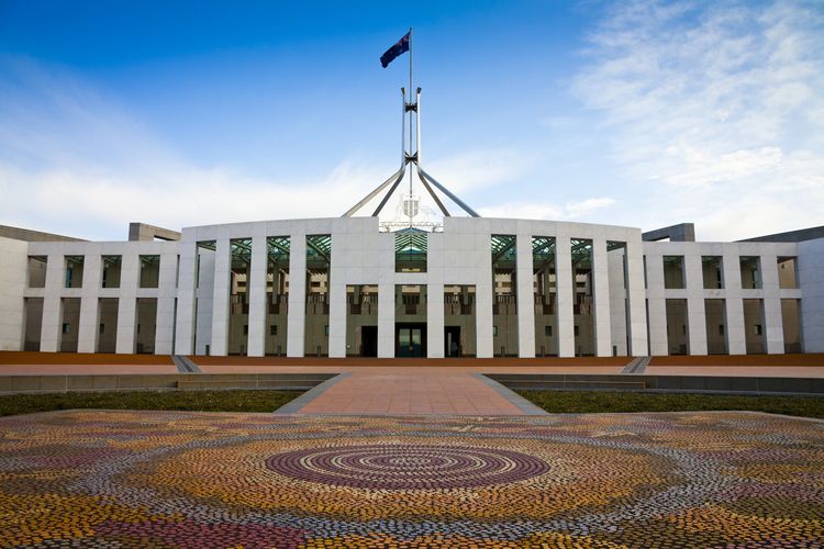 Australian Parliament House with flag and patterned forecourt under a clear sky.