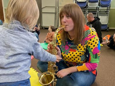 Jen playing saxophone while a toddler pushes the keys. With other children playing in the background