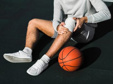Basketball player sitting with a knee injury, holding a bandaged knee beside a basketball.