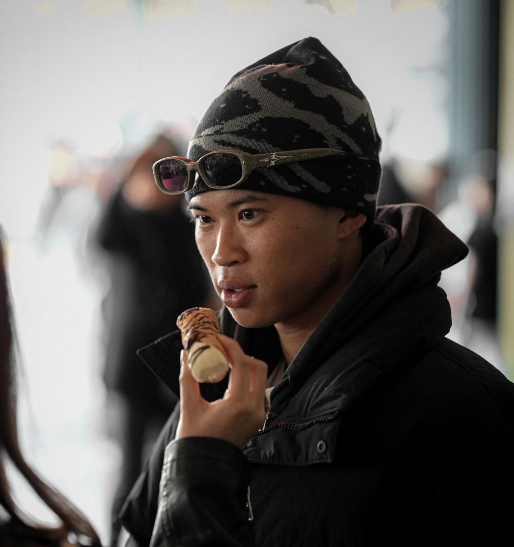 A man in a beanie and jacket looks at a small dessert held by another person.