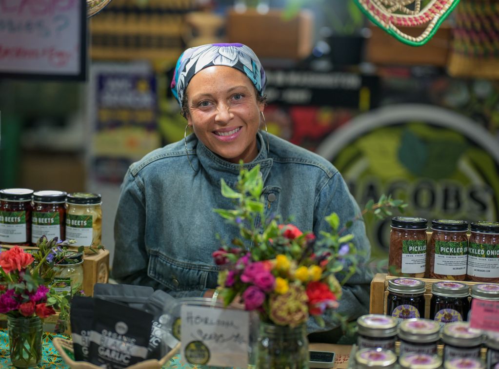 Smiling vendor at a market stall with colorful flowers and jars of pickled goods.