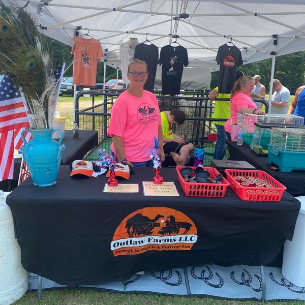 Woman in pink shirt at Outlaw Farms LLC booth with merchandise and trophies.