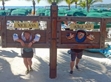 Two people posing in stocks labeled 'Nice Ass' and 'Dumb Ass' at a sunny beach.