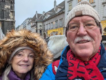A smiling elderly couple dressed warmly outdoors in a festive town setting.