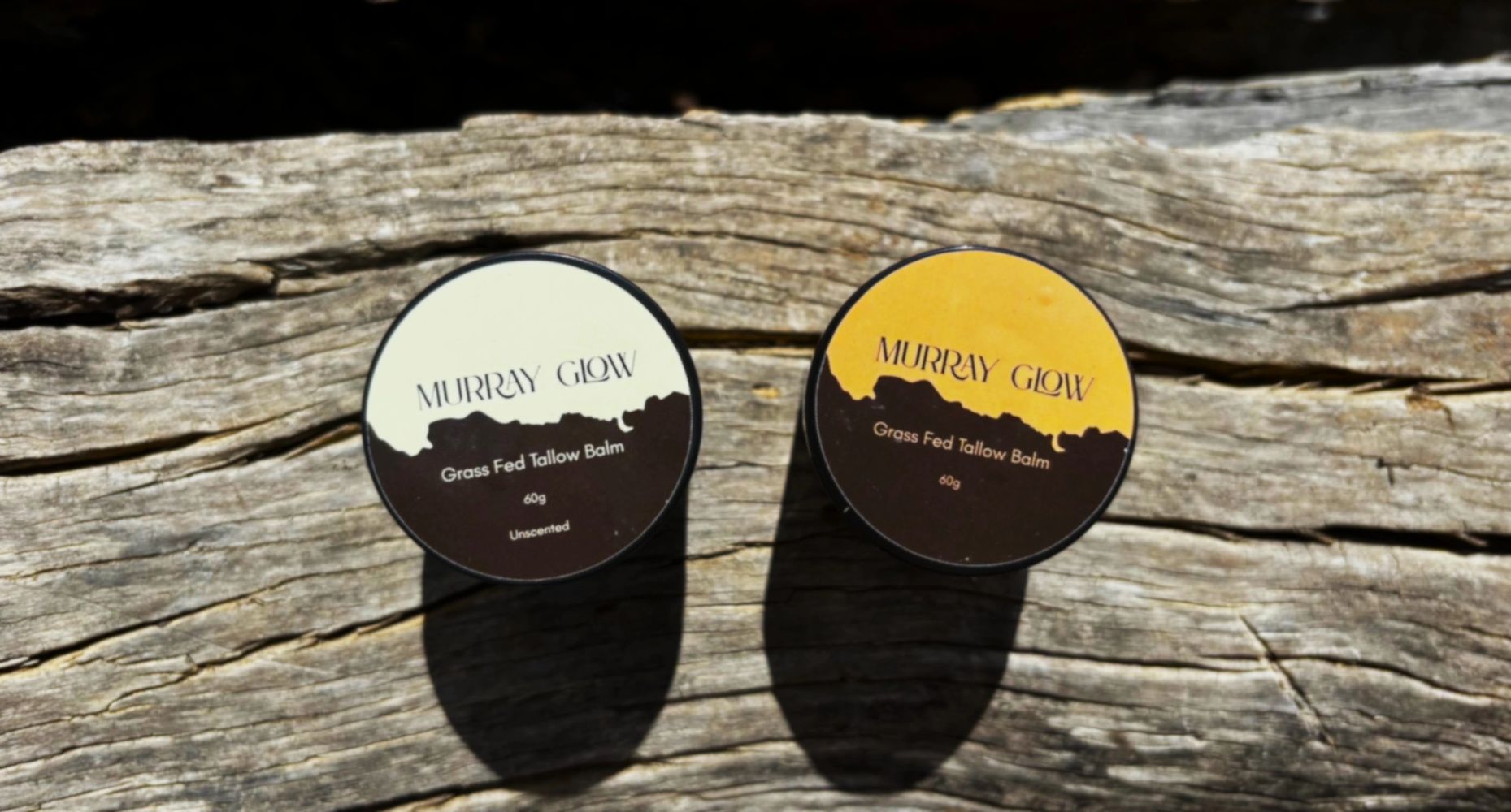 Two tins of Murray Glow Grass Fed Tallow Balm placed on rustic wood.