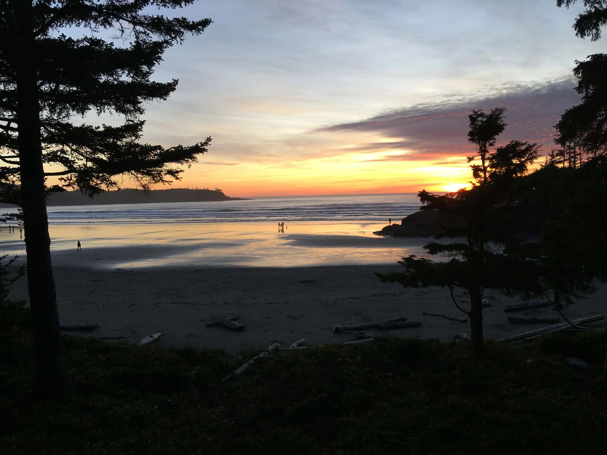 Sunset over a tranquil beach with silhouetted trees and people.
