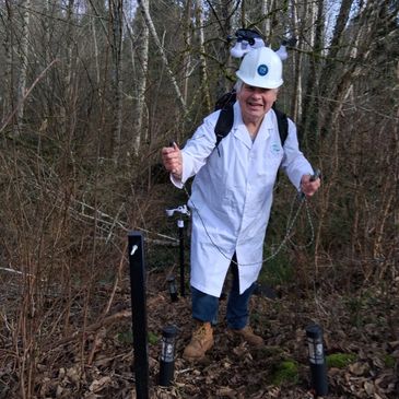 Scientist in lab coat and helmet exploring a wooded area with instruments.