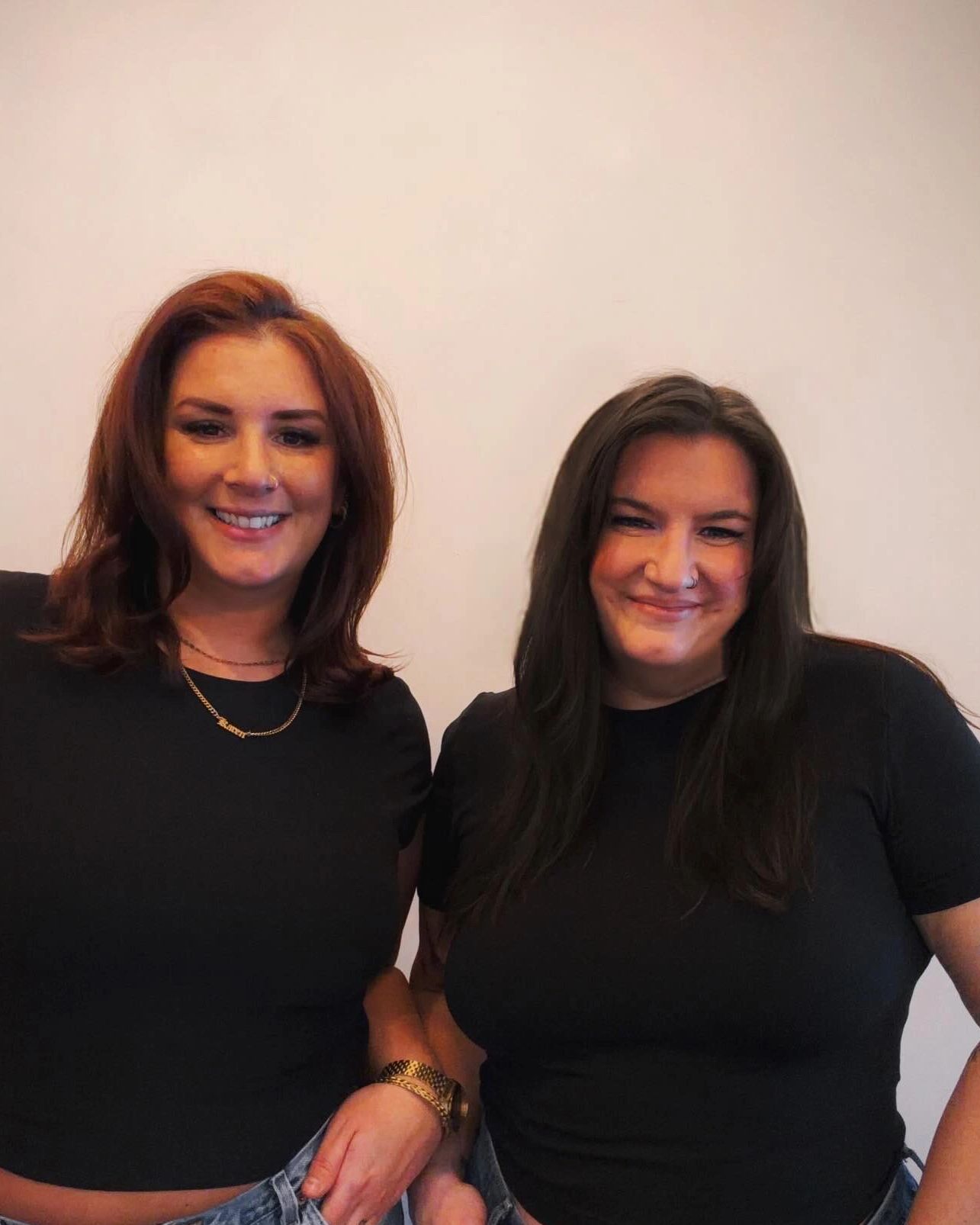 Two women smiling, dressed in black tops and jeans against a plain background.