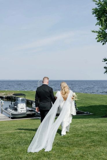 Bride and groom walking on grass near water with boats.