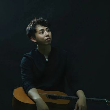 Young man with guitar in low light, looking thoughtfully.