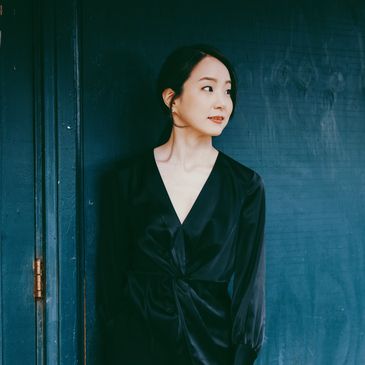 Woman in a black dress standing against a dark blue wall, looking to the side.