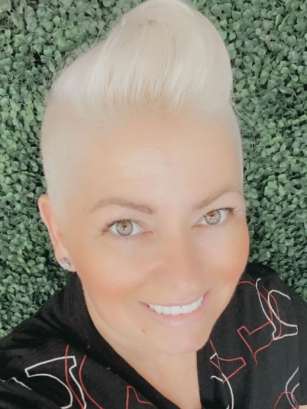 Smiling woman with platinum blonde hair styled upwards against a leafy green background.