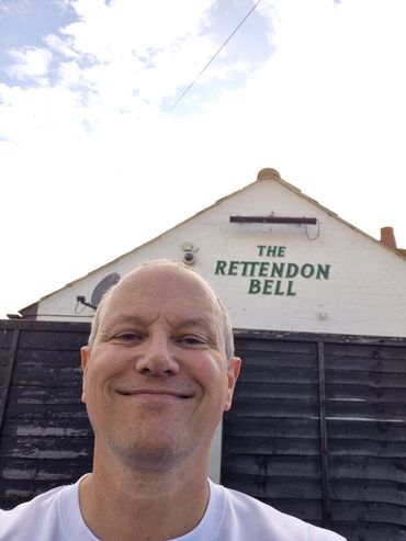 Smiling man taking a selfie outside The Rettendon Bell pub under a partly cloudy sky.