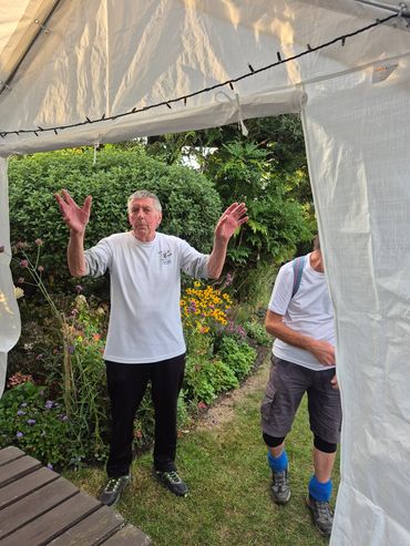 Two men outdoors with one gesturing, surrounded by greenery.