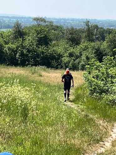 A person walking on a narrow path through a grassy field with trees in the background.