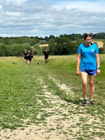 A woman in blue walking on a grassy path with others and a dog behind her.