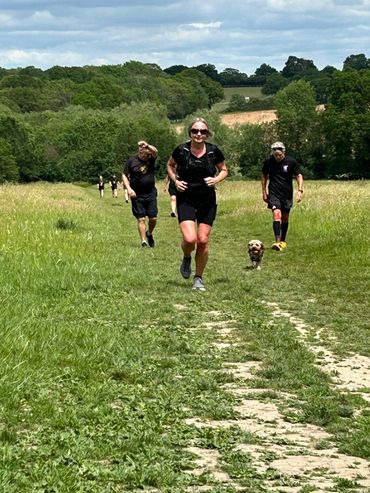 People jogging on a grassy trail with a small dog on a sunny day.