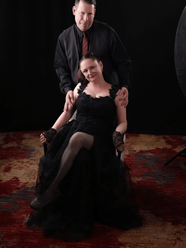 A couple posing elegantly with the woman seated in a black dress and the man standing behind her.