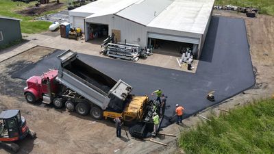 Construction workers paving a driveway near a warehouse with heavy machinery.
