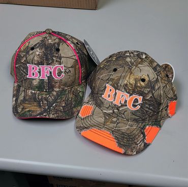 Two camouflage BFC caps with pink and orange accents on a gray surface.