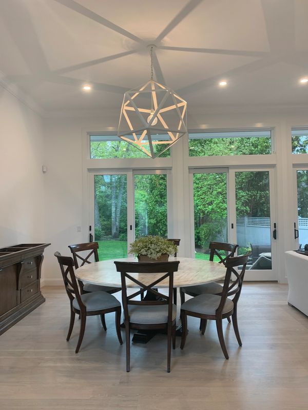 Modern dining room with wooden chairs, round table, and geometric chandelier by large windows.