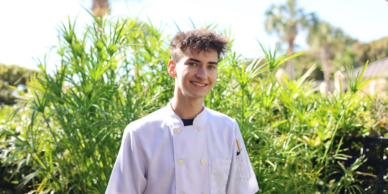 Young chef smiling outdoors in white uniform.
