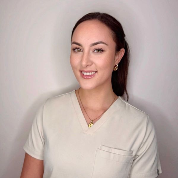 Smiling woman in light beige scrubs with gold jewelry.