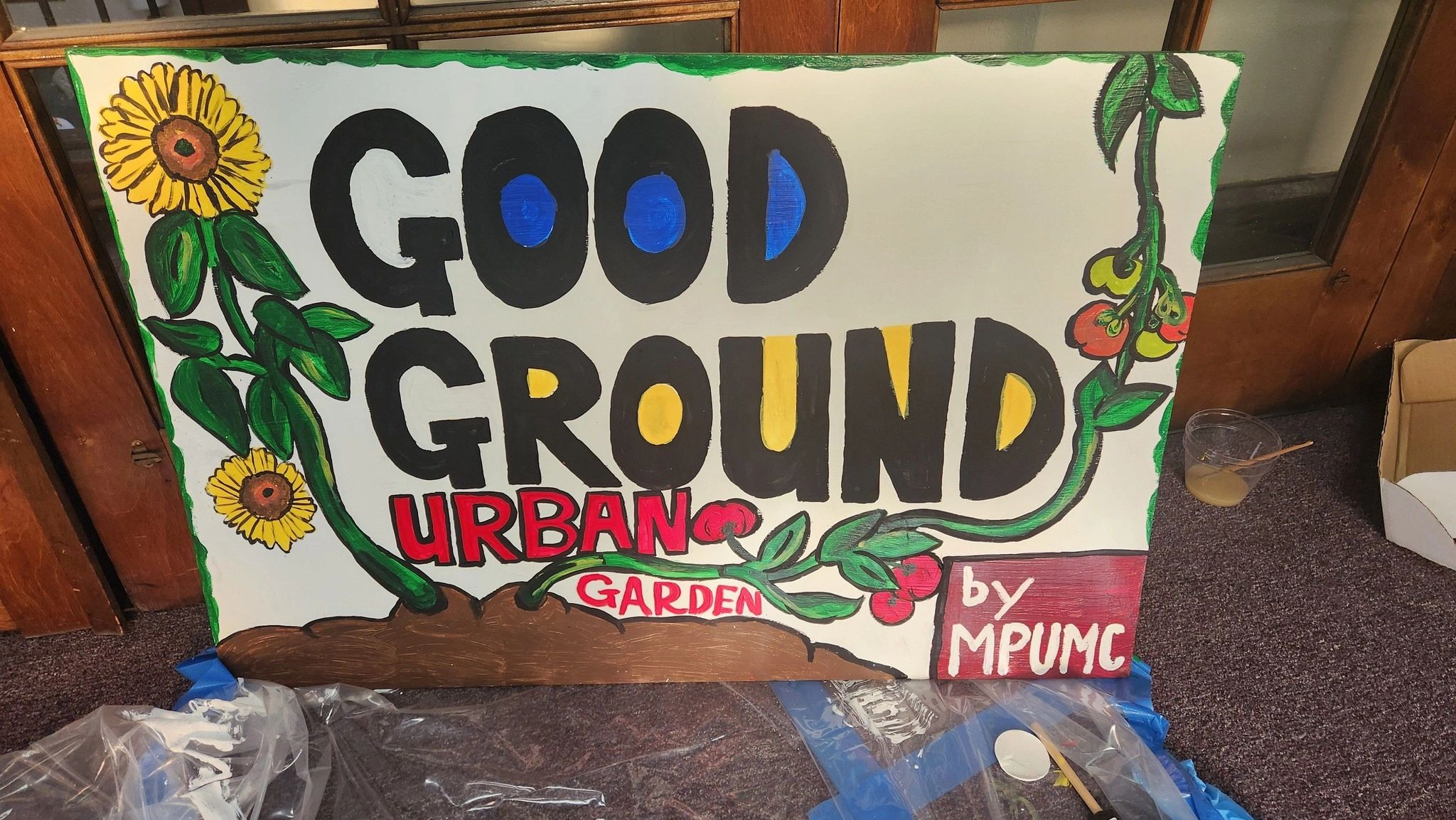 Bright handmade sign reading 'Good Ground Urban Garden by MPUMC' with floral decorations.