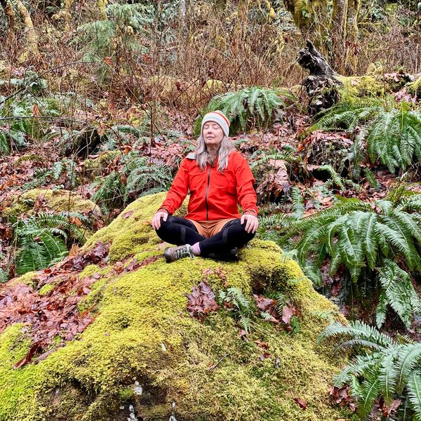 A woman meditates on a moss-covered rock in a lush forest.
